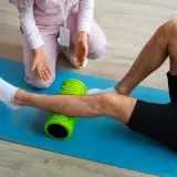 Un uomo esegue esercizi di stiramento polpaccio con un foam roller verde, assistito da un terapista.