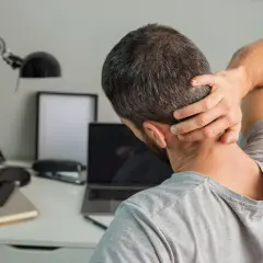 Uomo con mano sul collo, soffre di cefalea cervicogenica. Sulla scrivania, laptop, lampada e quaderni.