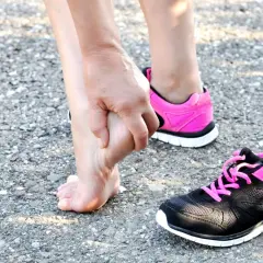 Donna si tocca il piede, forse per una contusione al calcagno. Tempi di recupero da valutare. Scarpe sportive rosa e nere.