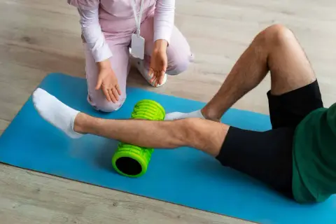 Un uomo esegue esercizi di stiramento polpaccio con un foam roller verde, assistito da un terapista.