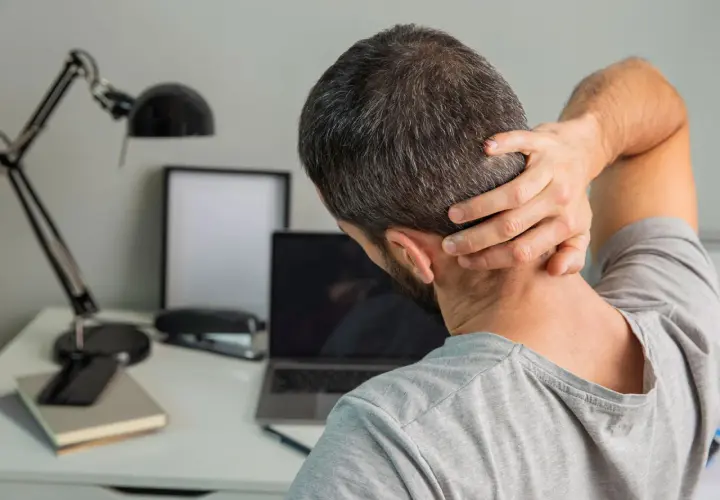 Uomo con mano sul collo, soffre di cefalea cervicogenica. Sulla scrivania, laptop, lampada e quaderni.