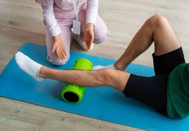 Un uomo esegue esercizi di stiramento polpaccio con un foam roller verde, assistito da un terapista.