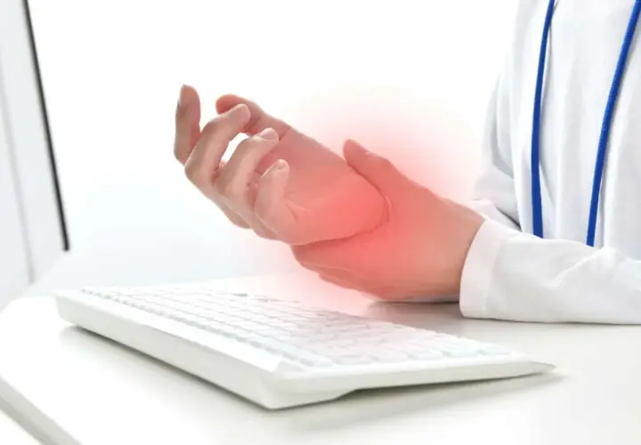 Mano che tiene il polso, con un'area rossa che indica dolore, vicino a una tastiera. Possibili tendiniti diffuse dovute all'uso prolungato del computer.