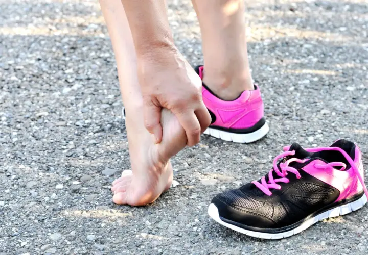 Donna si tocca il piede, forse per una contusione al calcagno. Tempi di recupero da valutare. Scarpe sportive rosa e nere.