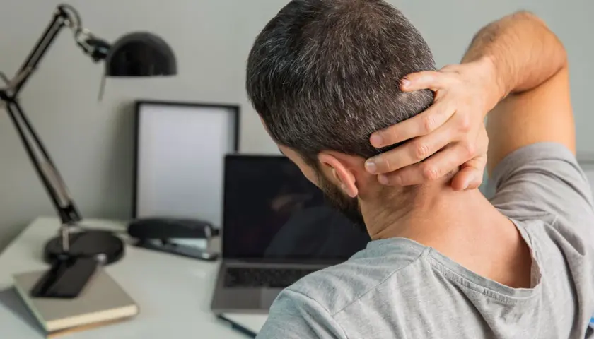 Uomo con mano sul collo, soffre di cefalea cervicogenica. Sulla scrivania, laptop, lampada e quaderni.