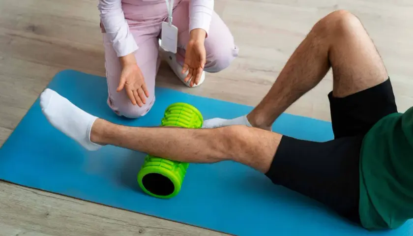 Un uomo esegue esercizi di stiramento polpaccio con un foam roller verde, assistito da un terapista.