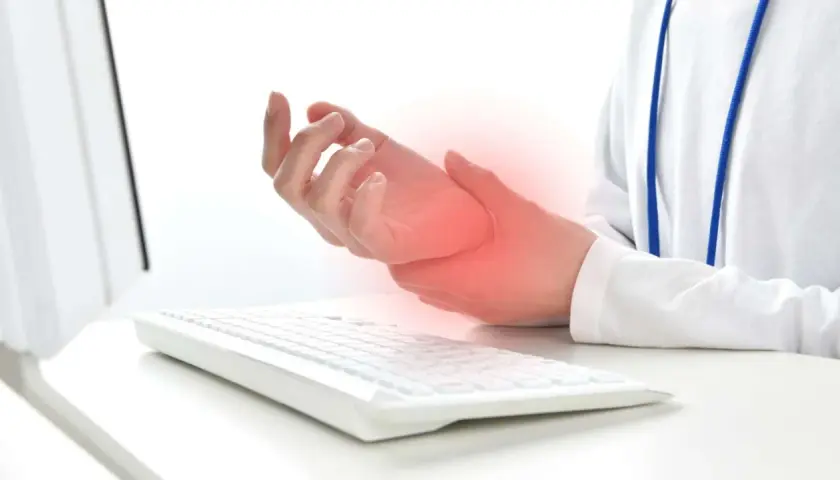 Mano che tiene il polso, con un'area rossa che indica dolore, vicino a una tastiera. Possibili tendiniti diffuse dovute all'uso prolungato del computer.