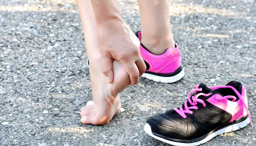 Donna si tocca il piede, forse per una contusione al calcagno. Tempi di recupero da valutare. Scarpe sportive rosa e nere.