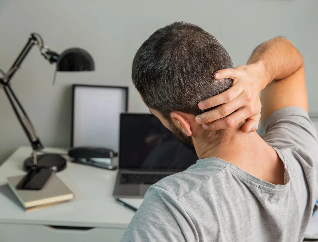 Uomo con mano sul collo, soffre di cefalea cervicogenica. Sulla scrivania, laptop, lampada e quaderni.