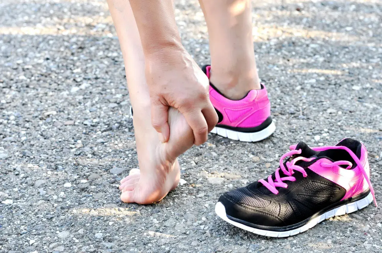 Donna si tocca il piede, forse per una contusione al calcagno. Tempi di recupero da valutare. Scarpe sportive rosa e nere.