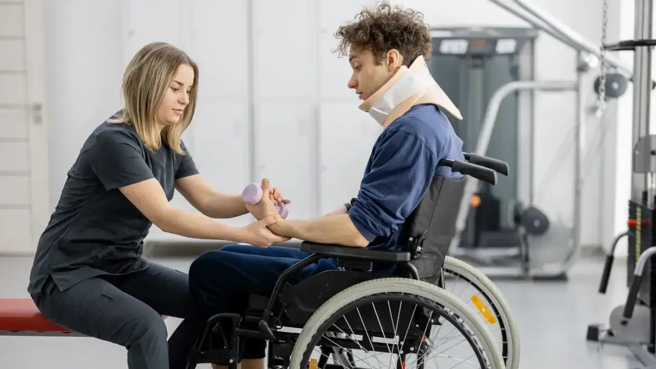 Fisioterapista assiste un paziente in sedia a rotelle con collare cervicale durante un esercizio per il controllo del tronco, usando un piccolo peso.
